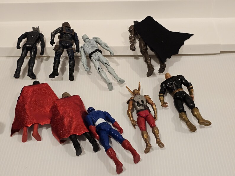 May include: A collection of superhero action figures in various poses. The figures are in different colors, including black, blue, red, gold, and gray. Some figures have capes. The figures are on a white surface.
