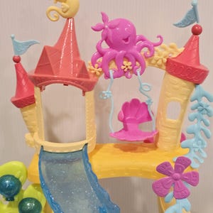 K&ouml;nnte beinhalten: Ein farbenfrohes Spielset aus Kunststoff, das einem Unterwasserschloss &auml;hnelt. Das Set verf&uuml;gt &uuml;ber eine Rutsche, T&uuml;rme mit Fahnen und dekorative Elemente wie ein Seepferdchen, einen Oktopus und eine Schaukel. Die Farbpalette umfasst Rosa, Gelb, Blau und Gr&uuml;n.