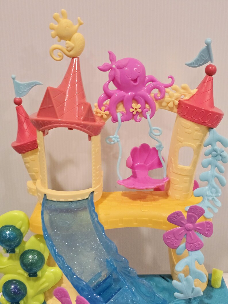K&ouml;nnte beinhalten: Ein farbenfrohes Spielset aus Kunststoff, das einem Unterwasserschloss &auml;hnelt. Das Set verf&uuml;gt &uuml;ber eine Rutsche, T&uuml;rme mit Fahnen und dekorative Elemente wie ein Seepferdchen, einen Oktopus und eine Schaukel. Die Farbpalette umfasst Rosa, Gelb, Blau und Gr&uuml;n.