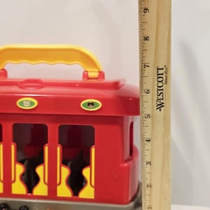 K&ouml;nnte beinhalten: Ein roter und gelber Spielzeug-Trolley mit Griff, mit grauem Sockel und gelben Akzenten. Der Trolley ist etwa 13 cm hoch. Zwei kleine Charakterfiguren sind im Vordergrund zu sehen.