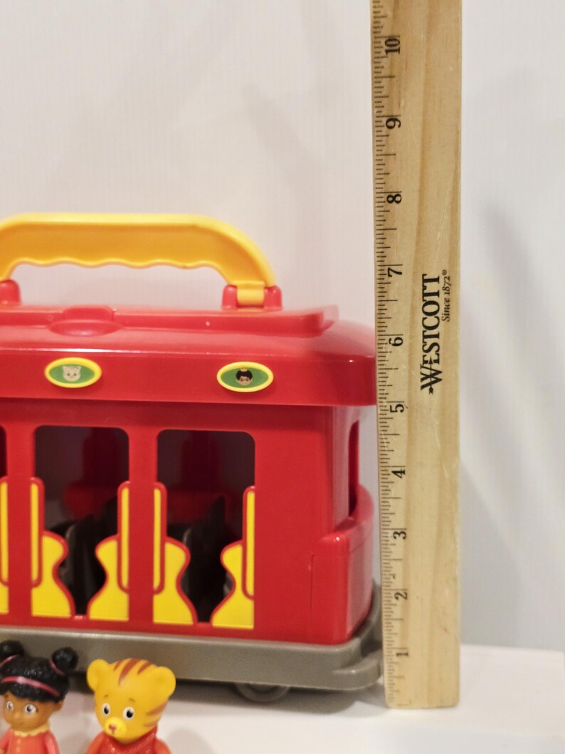 K&ouml;nnte beinhalten: Ein roter und gelber Spielzeug-Trolley mit Griff, mit grauem Sockel und gelben Akzenten. Der Trolley ist etwa 13 cm hoch. Zwei kleine Charakterfiguren sind im Vordergrund zu sehen.