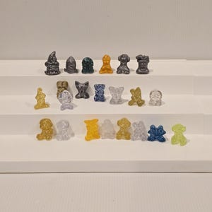 May include: A collection of small, colorful, and glittery resin figurines. The figurines are arranged on a white shelf and include various shapes such as dogs, bears, and abstract forms. Colors include gold, blue, green, and clear.