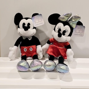以下が含まれることがあります： ミッキーマウスとミニーマウスのぬいぐるみが、クラシックな赤と黒の衣装を着ています。両方のぬいぐるみは、ホログラフィックデザインの銀色の靴を履いており、「Disney 100」とそれぞれの名前が書かれたタグが付いています。
