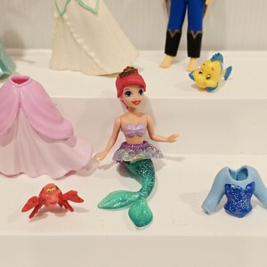 K&ouml;nnte beinhalten: Eine Sammlung farbenfroher Figuren mit Charakteren aus einem beliebten Animationsfilm. Die Figuren umfassen eine Meerjungfrau mit gr&uuml;nem Schwanz, eine rote Krabbe und andere Charaktere. Die Figuren befinden sich auf einer wei&szlig;en Oberfl&auml;che.