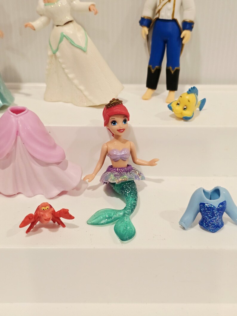 K&ouml;nnte beinhalten: Eine Sammlung farbenfroher Figuren mit Charakteren aus einem beliebten Animationsfilm. Die Figuren umfassen eine Meerjungfrau mit gr&uuml;nem Schwanz, eine rote Krabbe und andere Charaktere. Die Figuren befinden sich auf einer wei&szlig;en Oberfl&auml;che.