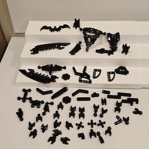 May include: A collection of black plastic building toy pieces, including various shapes like bat wings, gears, and connectors, arranged on white tiered platforms and a white surface. The pieces are of different sizes and shapes.