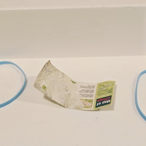 May include: A small, folded map with the text "Map of Village" in green and white. The map is slightly crumpled and rests on a white surface. Two blue rubber bands are visible on either side of the map.