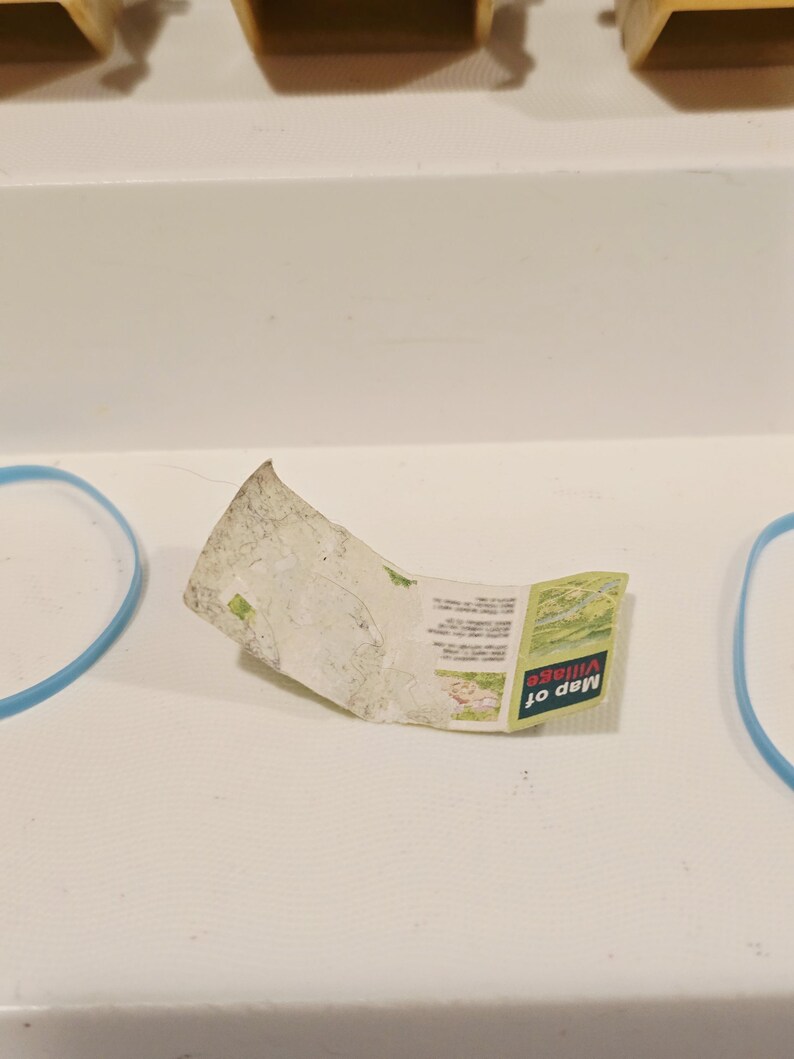 May include: A small, folded map with the text "Map of Village" in green and white. The map is slightly crumpled and rests on a white surface. Two blue rubber bands are visible on either side of the map.