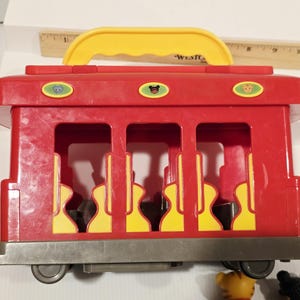 K&ouml;nnte beinhalten: Ein roter Spielzeug-Trolley mit gelbem Griff und drei ovalen Kn&ouml;pfen oben. Der Trolley hat drei offene Fenster mit gelb-schwarzen Sitzmustern. Der Trolley steht auf einer grauen Basis mit R&auml;dern.