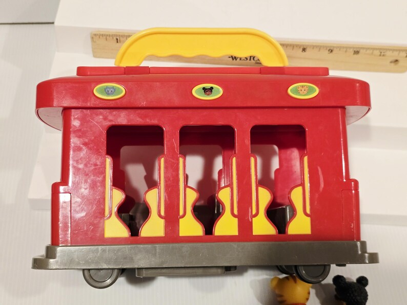 K&ouml;nnte beinhalten: Ein roter Spielzeug-Trolley mit gelbem Griff und drei ovalen Kn&ouml;pfen oben. Der Trolley hat drei offene Fenster mit gelb-schwarzen Sitzmustern. Der Trolley steht auf einer grauen Basis mit R&auml;dern.