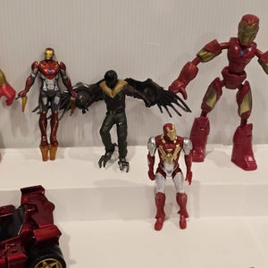 May include: A collection of action figures featuring characters from the Marvel universe. The figures include Iron Man in various poses, a Vulture figure with large black wings, and a red vehicle with gold wheels. The figures are displayed on a white shelf.