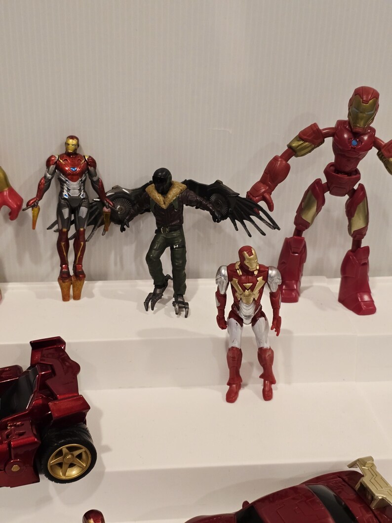 May include: A collection of action figures featuring characters from the Marvel universe. The figures include Iron Man in various poses, a Vulture figure with large black wings, and a red vehicle with gold wheels. The figures are displayed on a white shelf.