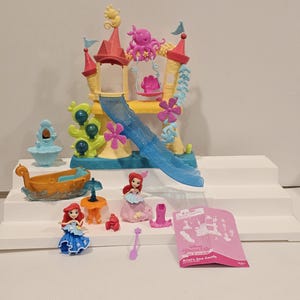 K&ouml;nnte beinhalten: Ariel's Sea Castle Spielset mit einem gelb-rosa Schloss mit einer blauen Rutsche, einem Boot und mehreren Ariel-Figuren. Das Set enth&auml;lt einen rosa Oktopus, einen Tisch und weiteres Zubeh&ouml;r. Die Verpackung tr&auml;gt die Aufschrift "Ariel's Sea Castle".