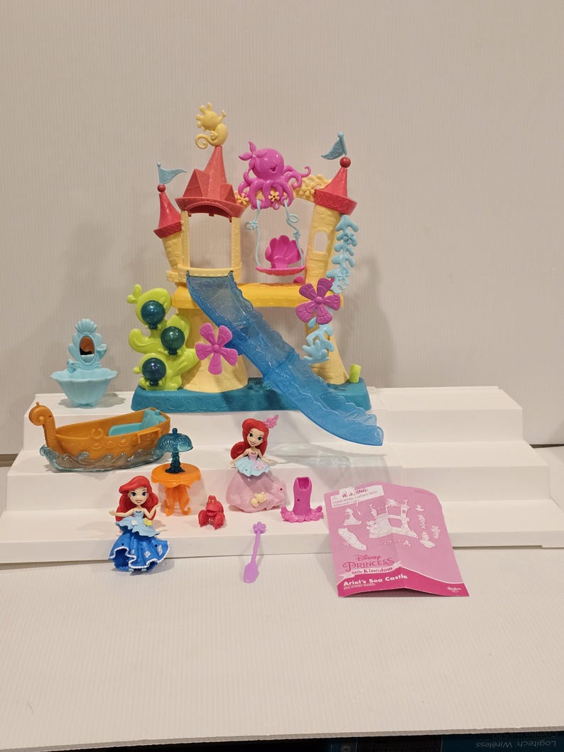 K&ouml;nnte beinhalten: Ariel's Sea Castle Spielset mit einem gelb-rosa Schloss mit einer blauen Rutsche, einem Boot und mehreren Ariel-Figuren. Das Set enth&auml;lt einen rosa Oktopus, einen Tisch und weiteres Zubeh&ouml;r. Die Verpackung tr&auml;gt die Aufschrift "Ariel's Sea Castle".