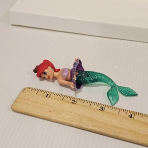 K&ouml;nnte beinhalten: Ariel-Puppenfigur mit rotem Haar, einem lila Oberteil und einem gr&uuml;nen Glitzerschwanz. Die Puppe liegt auf einer wei&szlig;en Oberfl&auml;che neben einem Holzlineal. Das Lineal misst etwa 10 cm.