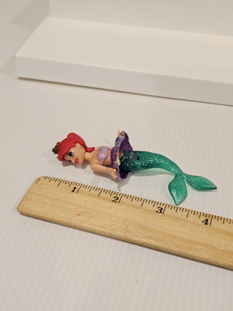 K&ouml;nnte beinhalten: Ariel-Puppenfigur mit rotem Haar, einem lila Oberteil und einem gr&uuml;nen Glitzerschwanz. Die Puppe liegt auf einer wei&szlig;en Oberfl&auml;che neben einem Holzlineal. Das Lineal misst etwa 10 cm.