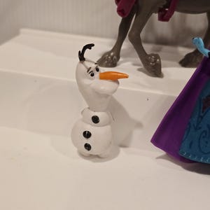 May include: A white Olaf figurine with a carrot nose, black hair, and three black buttons. The figurine is part of a larger set, with a reindeer and a purple cape visible in the background.
