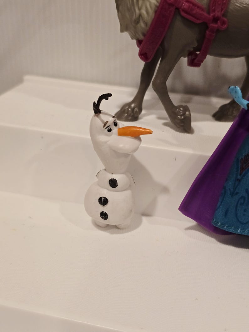 May include: A white Olaf figurine with a carrot nose, black hair, and three black buttons. The figurine is part of a larger set, with a reindeer and a purple cape visible in the background.