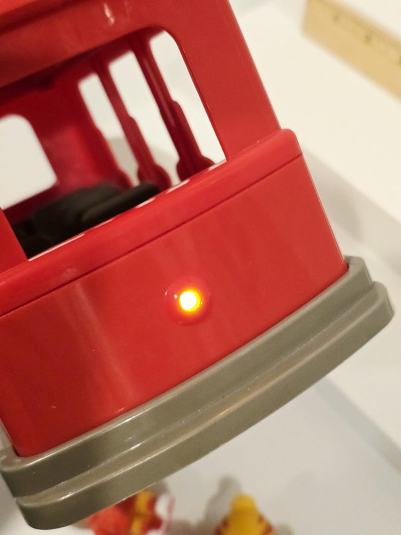 K&ouml;nnte beinhalten: Nahaufnahme eines roten Kunststoffspielzeugs mit grauem Sockel. Auf der Seite leuchtet ein kleines, rundes, gelbes Licht. Das Spielzeug hat eine rechteckige Form mit offenen R&auml;umen. Das Spielzeug steht auf einer wei&szlig;en Oberfl&auml;che.