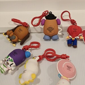 May include: A selection of Toy Story character keyrings. The keyrings feature characters such as Mr. Potato Head and Buzz Lightyear, each with a red clasp. The keyrings are made of a rubbery material and are brightly coloured. The keyrings are designed for attachment.