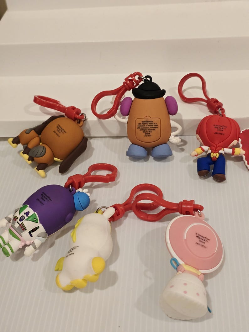May include: A selection of Toy Story character keyrings. The keyrings feature characters such as Mr. Potato Head and Buzz Lightyear, each with a red clasp. The keyrings are made of a rubbery material and are brightly coloured. The keyrings are designed for attachment.