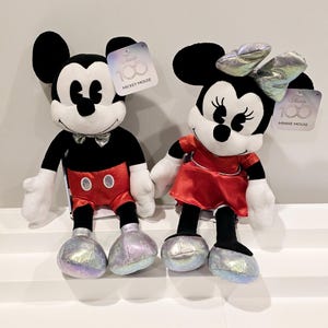 以下が含まれることがあります： ミッキーマウスとミニーマウスのぬいぐるみが、象徴的な赤と黒の衣装を着ています。白い表面に座っており、靴には銀色の輝きがあります。おもちゃのタグには、「Disney 100」、「ミッキーマウス」、「ミニーマウス」と書いてあります。