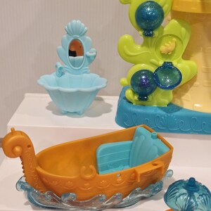 K&ouml;nnte beinhalten: Eine Sammlung farbenfroher Plastikspielzeuge, darunter ein kleines orangefarbenes Boot mit blauen Sitzen, ein hellblaues Waschbecken mit Spiegel und eine gelb-blaue burg&auml;hnliche Struktur. Das Boot steht auf einer durchscheinenden blauen Wellenbasis.