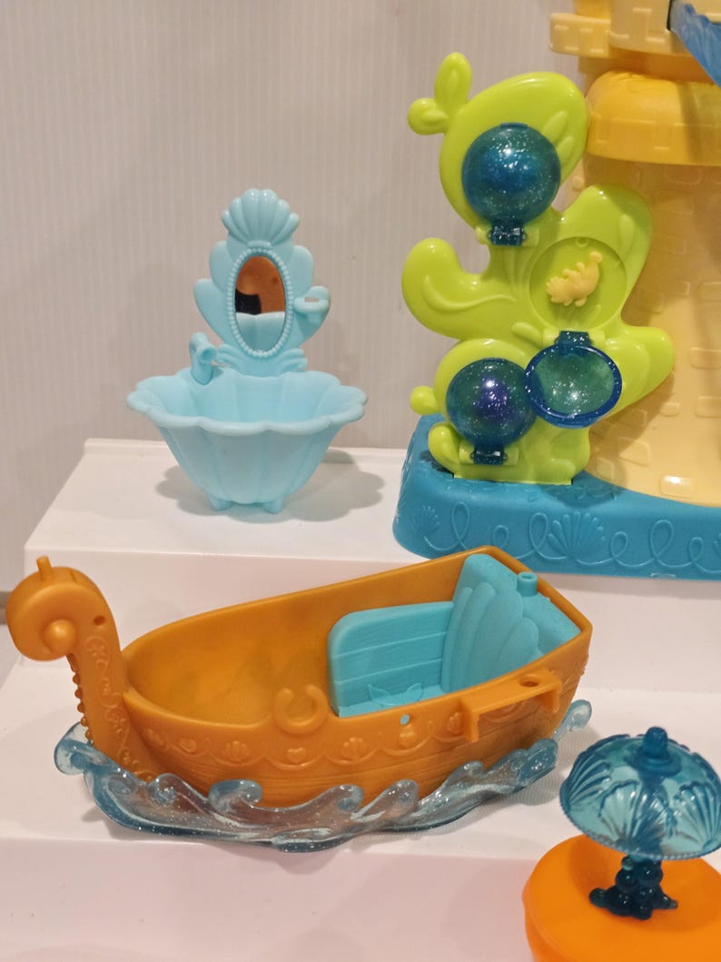 K&ouml;nnte beinhalten: Eine Sammlung farbenfroher Plastikspielzeuge, darunter ein kleines orangefarbenes Boot mit blauen Sitzen, ein hellblaues Waschbecken mit Spiegel und eine gelb-blaue burg&auml;hnliche Struktur. Das Boot steht auf einer durchscheinenden blauen Wellenbasis.