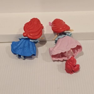 K&ouml;nnte beinhalten: Zwei kleine Prinzessinnenfiguren mit roten Haaren, eine in einem blauen Kleid und die andere in einem rosa Kleid. Eine kleine rote Krabbenfigur ist ebenfalls vorhanden. Die Figuren stehen auf einer wei&szlig;en Oberfl&auml;che.
