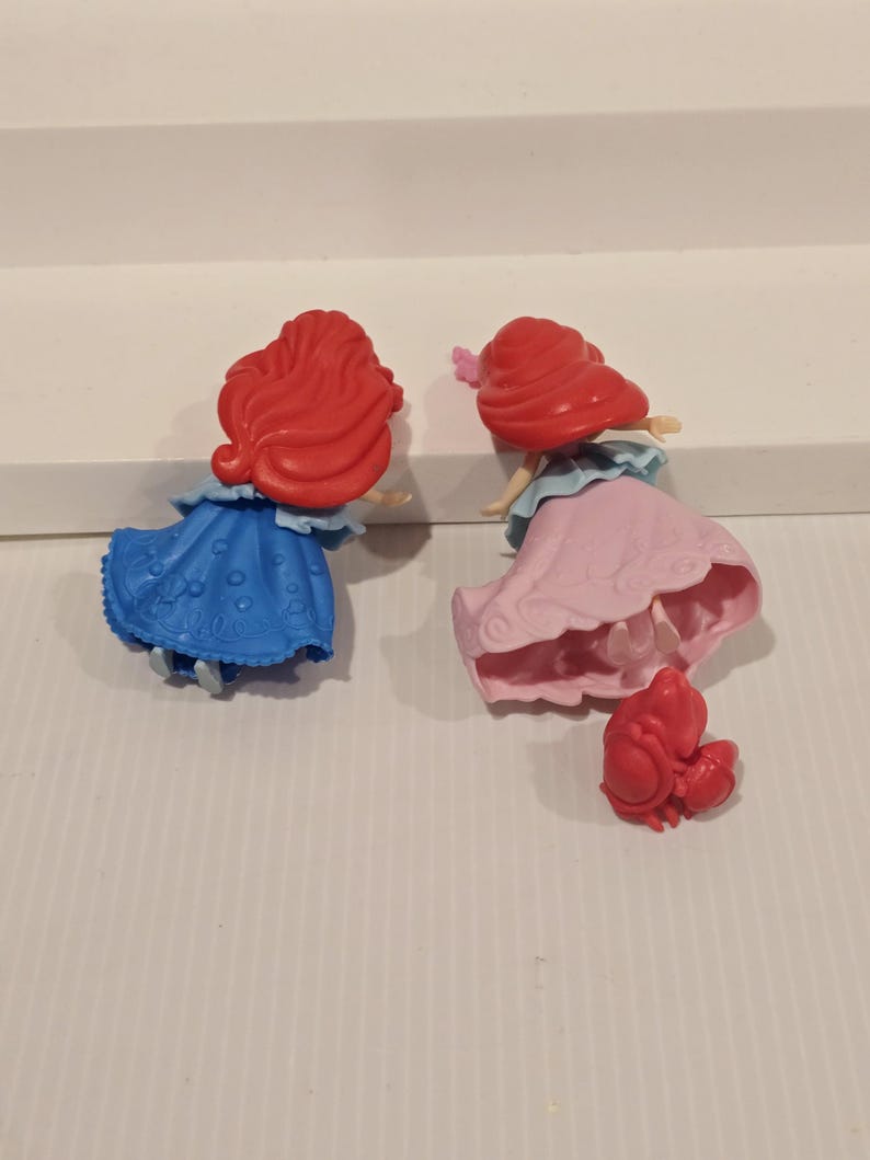 K&ouml;nnte beinhalten: Zwei kleine Prinzessinnenfiguren mit roten Haaren, eine in einem blauen Kleid und die andere in einem rosa Kleid. Eine kleine rote Krabbenfigur ist ebenfalls vorhanden. Die Figuren stehen auf einer wei&szlig;en Oberfl&auml;che.