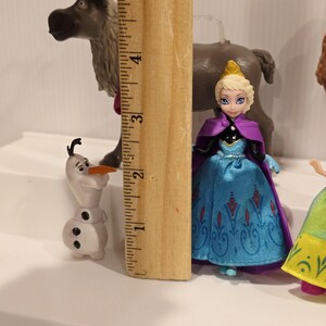 May include: A collection of Frozen character figurines, including Elsa in a blue dress with a purple cape, Olaf the snowman, and Sven the reindeer. The ruler shows Elsa is approximately 3 inches tall.