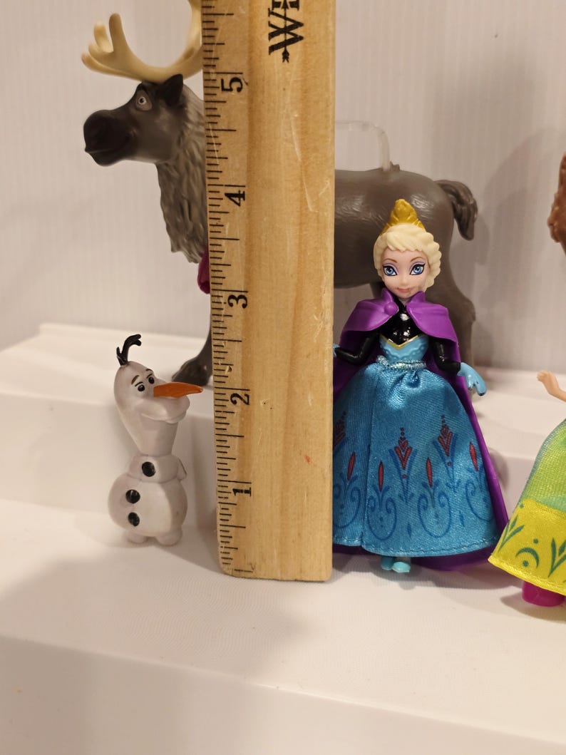 May include: A collection of Frozen character figurines, including Elsa in a blue dress with a purple cape, Olaf the snowman, and Sven the reindeer. The ruler shows Elsa is approximately 3 inches tall.