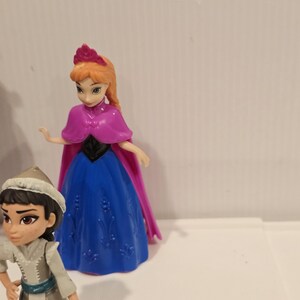 May include: Two plastic figurines of characters from the animated film "Frozen." One figure is dressed in a blue and purple gown with a pink cape, and the other is wearing a gray outfit with a white hat. Both figures have detailed facial features.