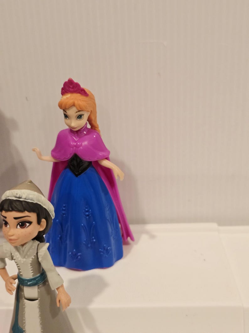May include: Two plastic figurines of characters from the animated film "Frozen." One figure is dressed in a blue and purple gown with a pink cape, and the other is wearing a gray outfit with a white hat. Both figures have detailed facial features.