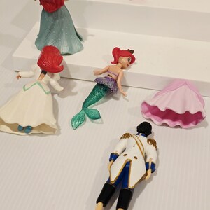 K&ouml;nnte beinhalten: Eine Sammlung kleiner Plastikfiguren mit Charakteren aus einem beliebten Zeichentrickfilm. Die Figuren umfassen eine Meerjungfrau mit gr&uuml;nem Schwanz, einen Prinzen in einer wei&szlig;en Jacke und verschiedene Meereskreaturen. Die Meerjungfrau hat rotes Haar und ein lila Oberteil.