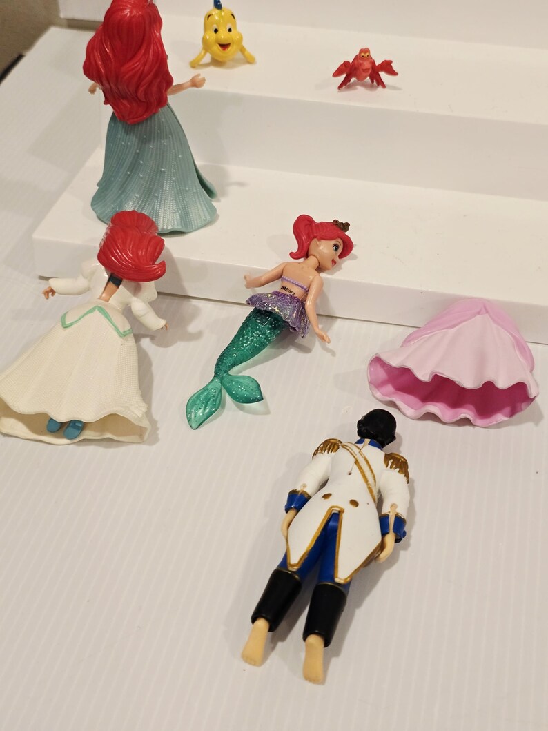 K&ouml;nnte beinhalten: Eine Sammlung kleiner Plastikfiguren mit Charakteren aus einem beliebten Zeichentrickfilm. Die Figuren umfassen eine Meerjungfrau mit gr&uuml;nem Schwanz, einen Prinzen in einer wei&szlig;en Jacke und verschiedene Meereskreaturen. Die Meerjungfrau hat rotes Haar und ein lila Oberteil.