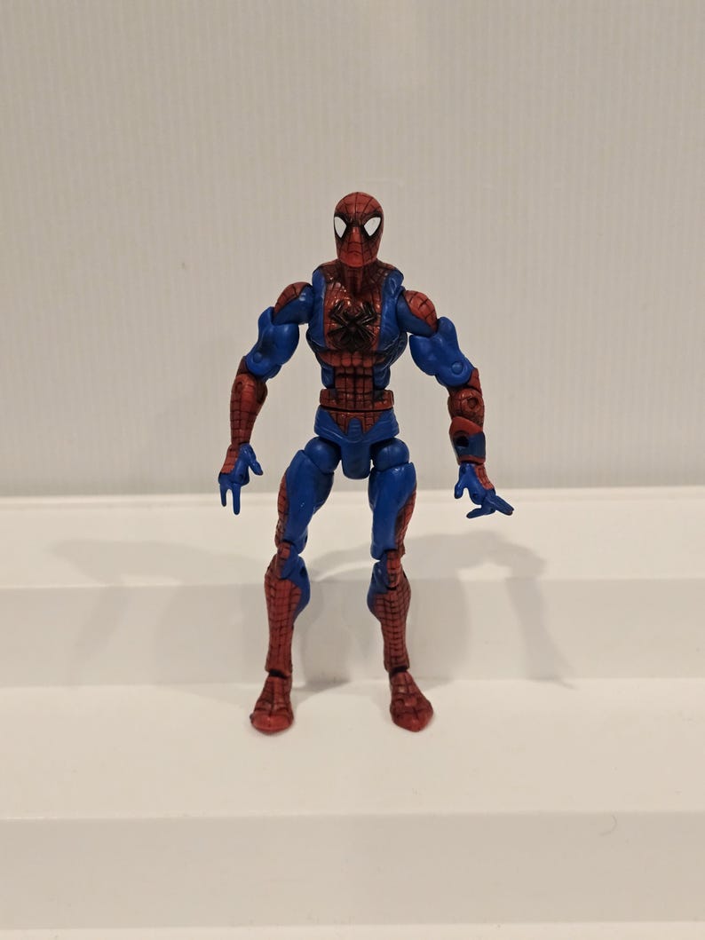 Pode incluir: Uma figura de a&ccedil;&atilde;o do Homem-Aranha em pose de p&eacute;. A figura &eacute; principalmente vermelha e azul, com um emblema de aranha preta no peito. Design detalhado, com m&uacute;sculos vis&iacute;veis e uma m&aacute;scara cl&aacute;ssica do Homem-Aranha.