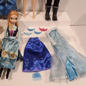 May include: A selection of dolls and accessories. There is a doll in a blue and gold dress, a blue skirt, and a light blue dress with a sheer overlay. Accessories include tiaras and a hair comb.