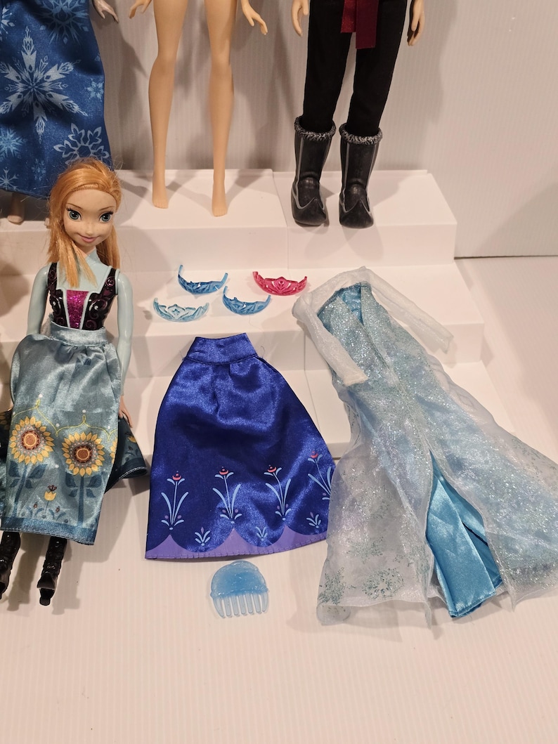 May include: A selection of dolls and accessories. There is a doll in a blue and gold dress, a blue skirt, and a light blue dress with a sheer overlay. Accessories include tiaras and a hair comb.