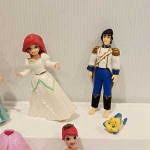 K&ouml;nnte beinhalten: Eine Sammlung von Disney-Figuren mit Charakteren aus Arielle, die Meerjungfrau. Die Figuren umfassen Arielle in einem wei&szlig;en Kleid, Prinz Eric in einer wei&szlig;-blauen Uniform und Fabius. Weitere Figuren sind ebenfalls sichtbar.