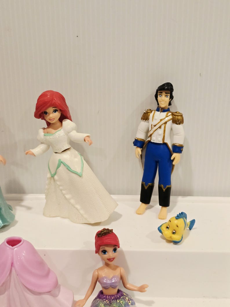 K&ouml;nnte beinhalten: Eine Sammlung von Disney-Figuren mit Charakteren aus Arielle, die Meerjungfrau. Die Figuren umfassen Arielle in einem wei&szlig;en Kleid, Prinz Eric in einer wei&szlig;-blauen Uniform und Fabius. Weitere Figuren sind ebenfalls sichtbar.