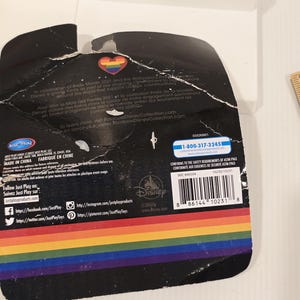 May include: A black product package with a rainbow-colored stripe at the bottom. The package has text and logos, including the Disney logo and a heart-shaped emblem with a rainbow design. The package is slightly torn.
