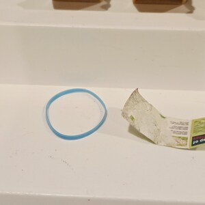 May include: A light blue rubber band and a torn piece of paper with green and white text are on a white surface. The paper appears to be a map. Several small, tan chairs are in the background.