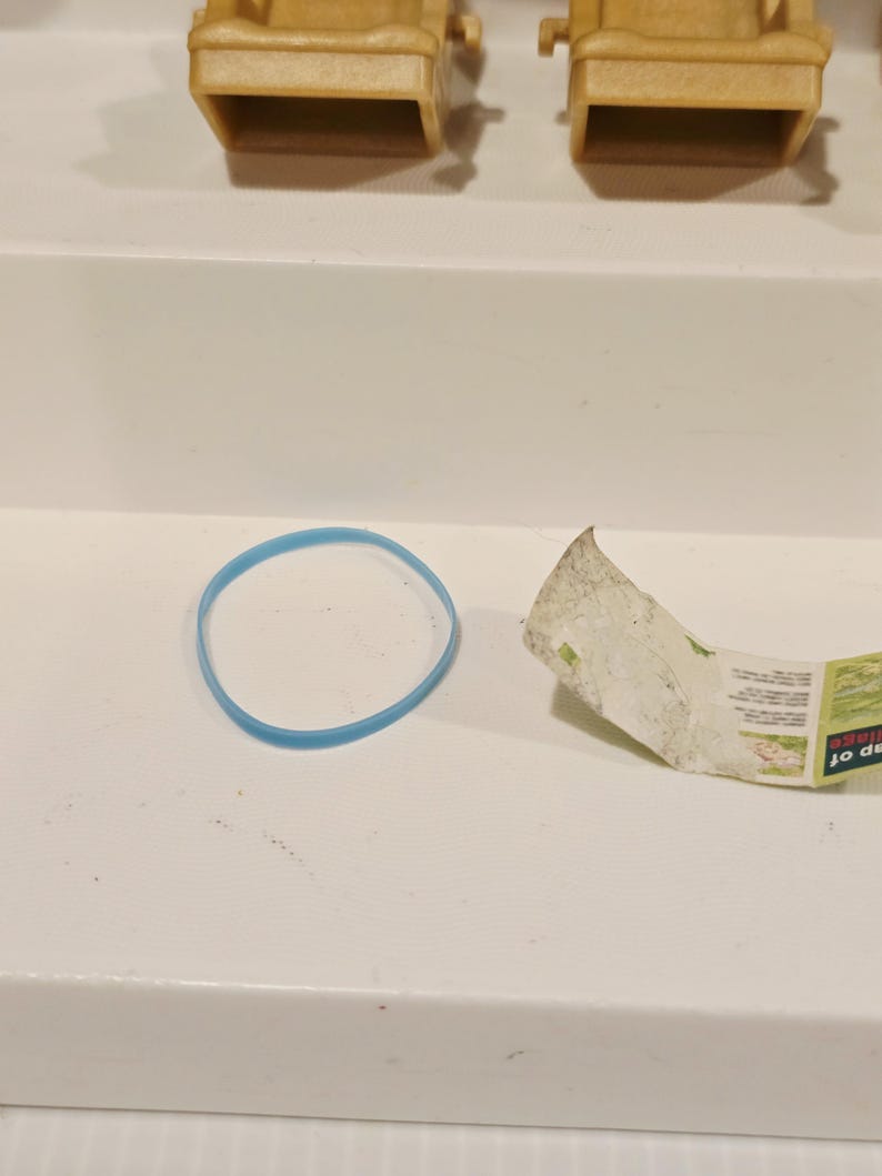 May include: A light blue rubber band and a torn piece of paper with green and white text are on a white surface. The paper appears to be a map. Several small, tan chairs are in the background.