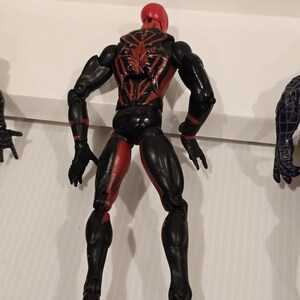 May include: A black and red action figure of a superhero, viewed from the back. The figure has a red head and accents, with a spider-like design on the back. The legs are red and black, and the figure is posed with arms at its sides.