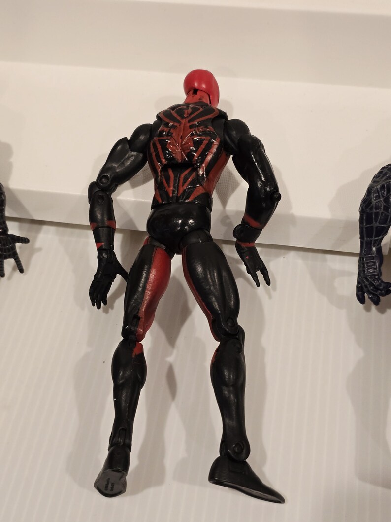 May include: A black and red action figure of a superhero, viewed from the back. The figure has a red head and accents, with a spider-like design on the back. The legs are red and black, and the figure is posed with arms at its sides.