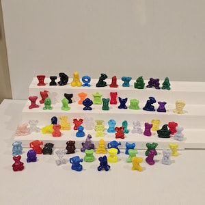 May include: A collection of small, colorful plastic figurines in various shapes, including animals and abstract forms. The figurines are arranged on a white surface and display a range of colors such as red, blue, yellow, and green. The collection appears to be a vintage toy set.