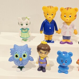 May include: A collection of six plastic character figurines including a white cat in a green dress, two orange tiger figures in blue and white clothing, a blue owl, a human figure with a crown, and a small blue bear-like creature.