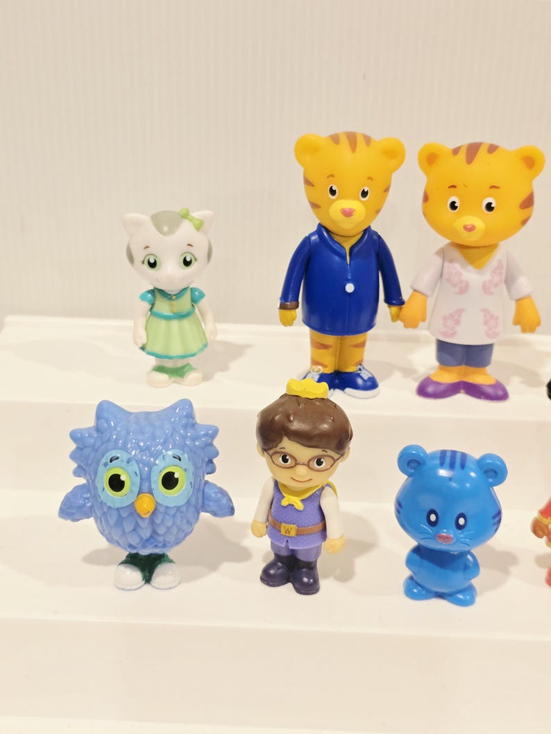 May include: A collection of six plastic character figurines including a white cat in a green dress, two orange tiger figures in blue and white clothing, a blue owl, a human figure with a crown, and a small blue bear-like creature.