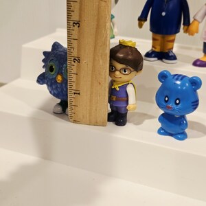 May include: A collection of small plastic character figurines including a blue owl, a figure with a gold crown, and a blue bear, positioned next to a wooden ruler showing a height of approximately 4 inches for the tallest visible figure.